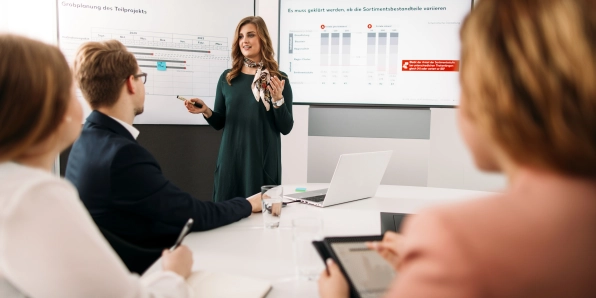 Eine Kaufland-Mitarbeiterin präsentiert im Konferenzraum. Im Vordergrund des Bildes sitzen ein Kollege und zwei Kolleginnen an einem ovalen Tisch und hören dem Vortrag ihrer Kollegin aufmerksam zu.
