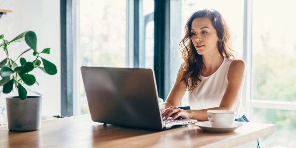 Frau sitzt daheim vor großen Fenstern und arbeitet an einem Laptop.