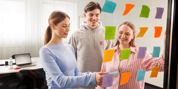 Zwei junge Frauen und ein junger Mann in legerer Kleidung stehen vor einer gläsernen Wand und arbeiten mit Post-Its an einem Projektboard.
