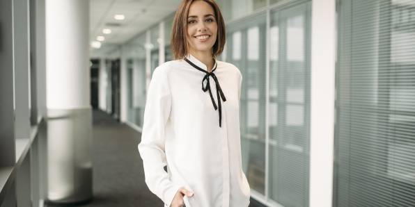 Unsere Teamleiterin für das Supply Chain Management bei Kaufland posiert lächelnd in weißer Bluse auf dem Büroflur.