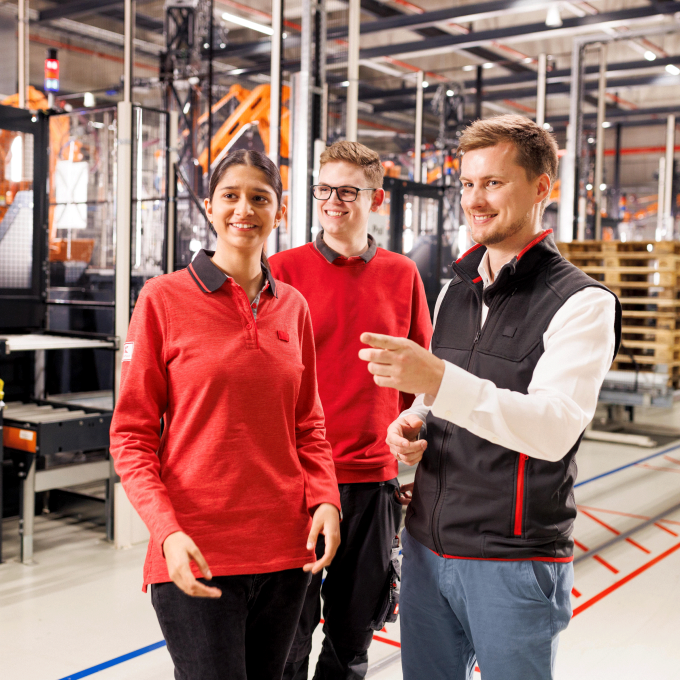 Ein junger Mitarbeiter, eine junge Mitarbeiterin, beide in rotem Pullover, sowie ein Ausbilder arbeiten in der Logistik. Der Ausbilder erklärt etwas.