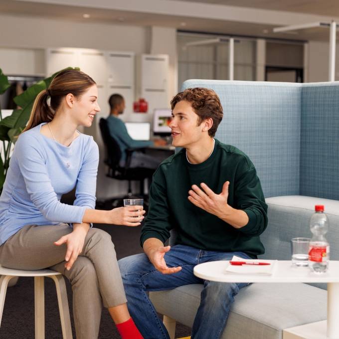 Eine Frau und ein junger Mann sitzen leger gekleidet auf einem Sofa und auf einem Stuhl. In der Erklärsituation zwischen Patin und Patenkind erläutert der Mann etwas.