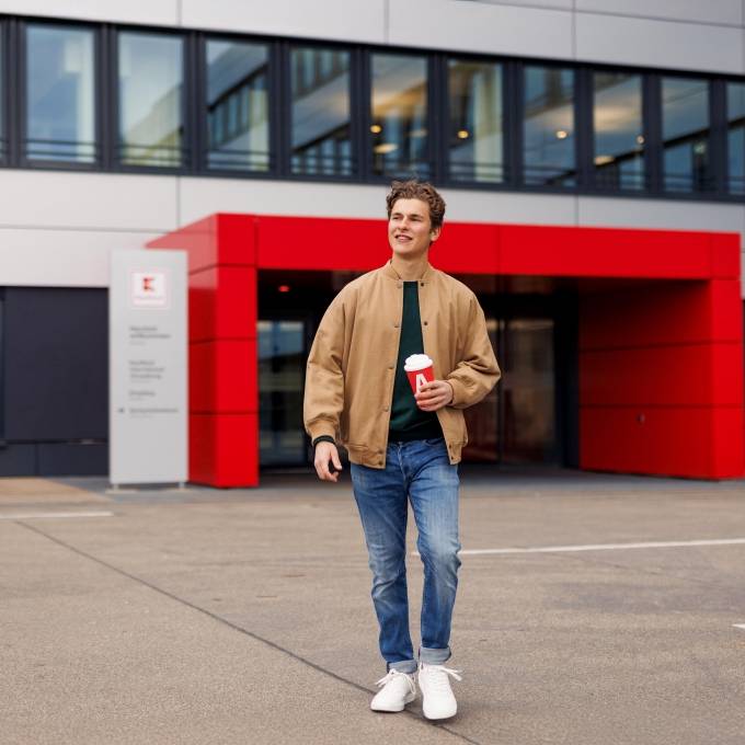 Ein Azubi, ein junger Mann in brauner Jacke, geht über den Platz vor dem Haupteingang der Kaufland-Zentrale.