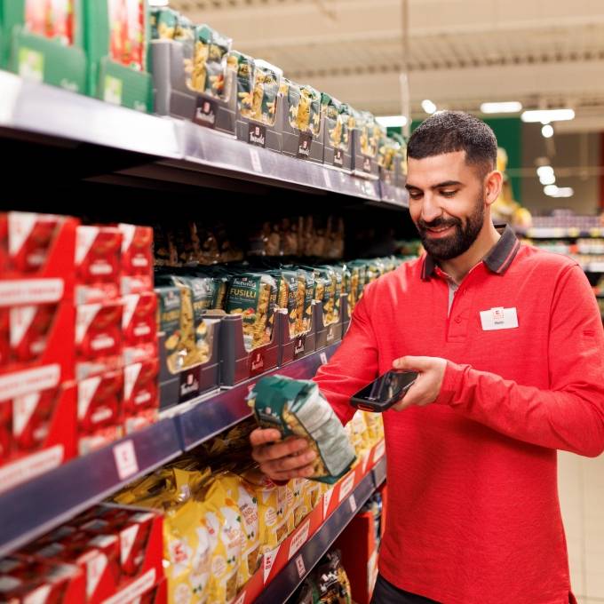 Ein Mitarbeiter in roter Kaufland-Kleidung steht in einem Gang und scannt eine Packung Nudeln.