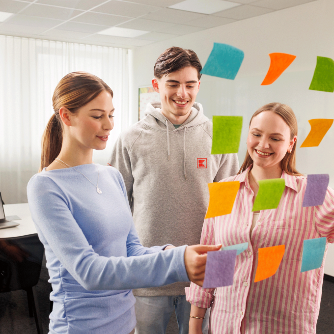 Zwei junge Frauen und ein junger Mann in legerer Kleidung stehen vor einer gläsernen Wand und arbeiten mit Post-Its an einem Projektboard.