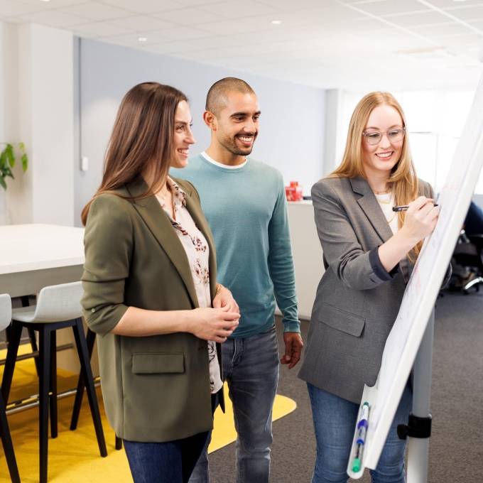 In der Workshopsituation stehen eine junge Frau und ein junger Mann, beide Trainees, mit einer Ausbilderin vor einem Flipchart.