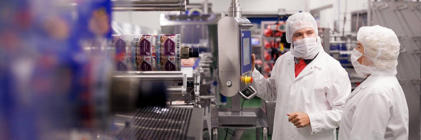 Fertigungsanlage in einem Kaufland-Fleischwerk. Zwei Kaufland-Mitarbeiter bedienen eine Produktionsanlage und unterhalten sich.