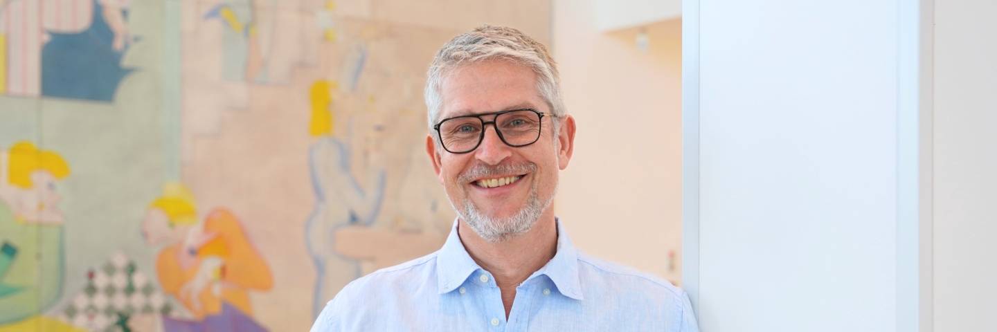 Porträt des lächelnden Managers Knut Harenkamp mit Brille und hellem Hemd, stehend vor einem modernen, farbigen Kunstwerk (Kaufland Karriere).