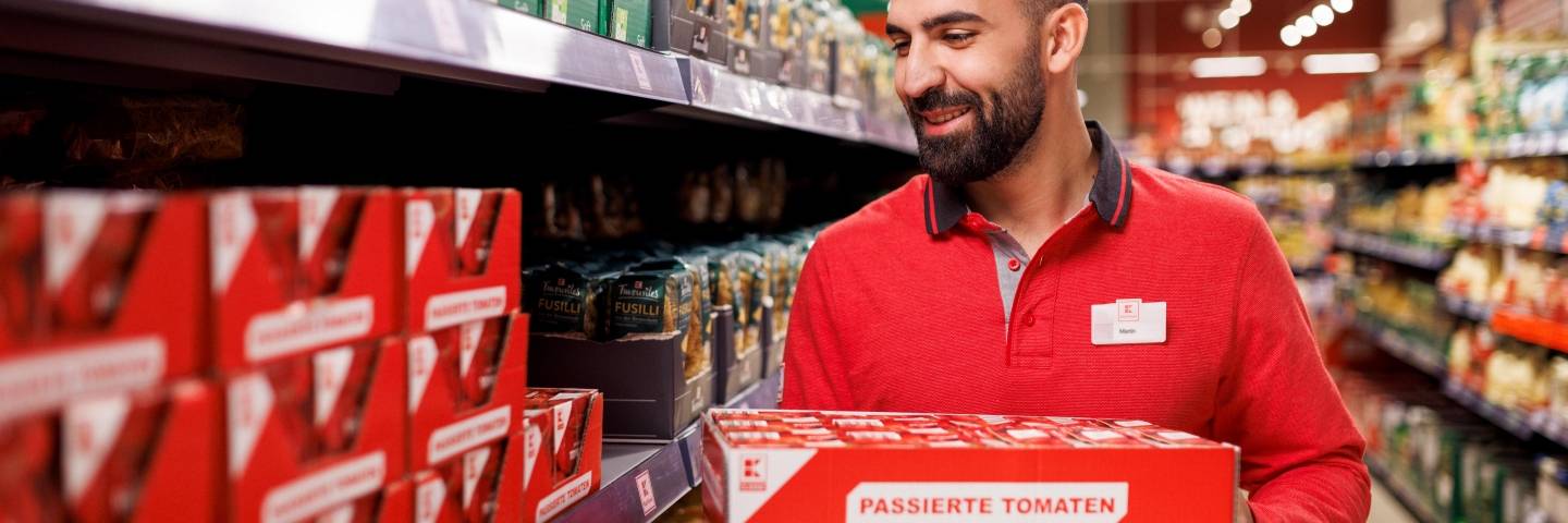 Ein Mitarbeiter in roter Kaufland-Kleidung steht in einem Gang, trägt einen Karton und verräumt Ware.
