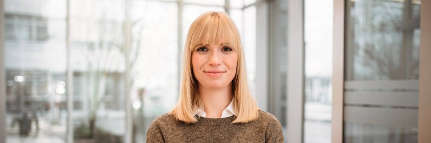 Eine Kaufland-Mitarbeiterin steht mit Laptop vor einer großen Glasfront und Sitzgruppe in einem lichtdurchfluteten, modernen Empfangsraum.