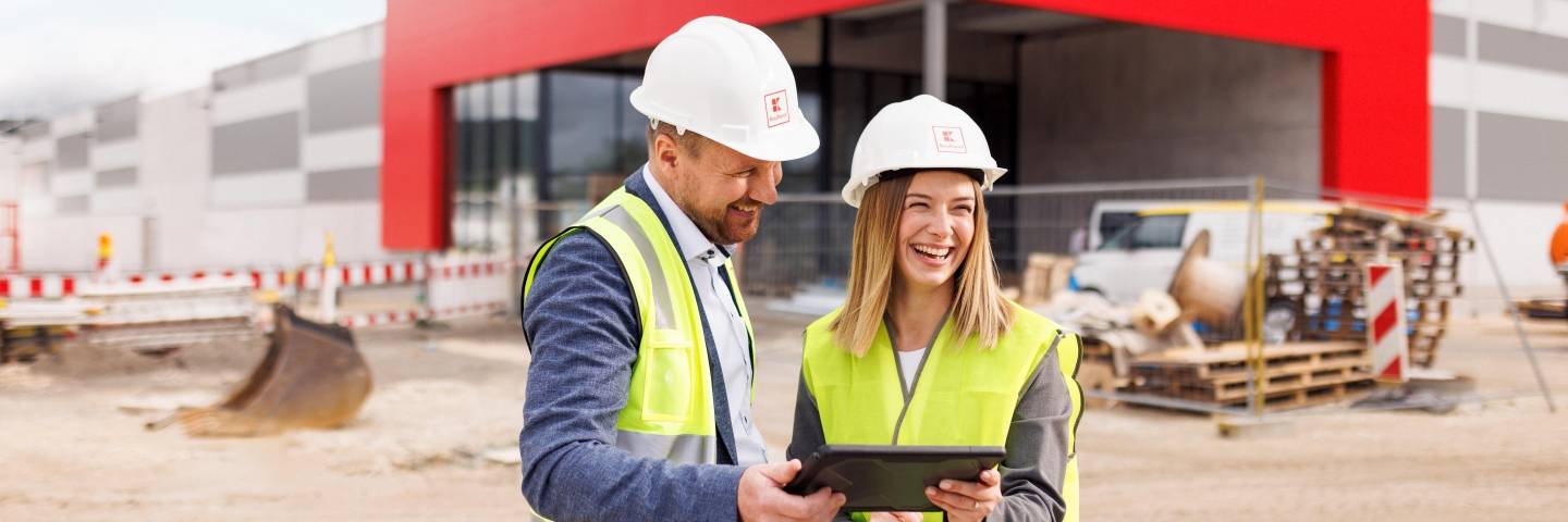 Zwei Mitarbeiter stehen vor dem Bau eines Kaufland-Marktes und halten ein Tablet in den Händen.