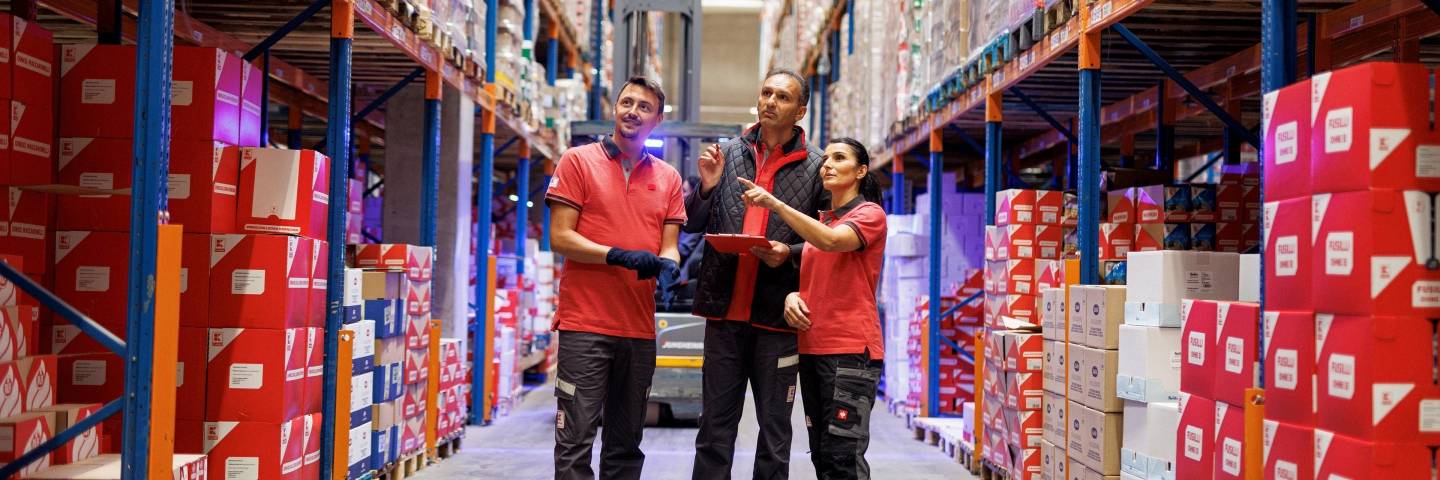 Drei Logistikmitarbeiter von Kaufland in einem Lagergang mit hohen, gut gefüllten Regalen.