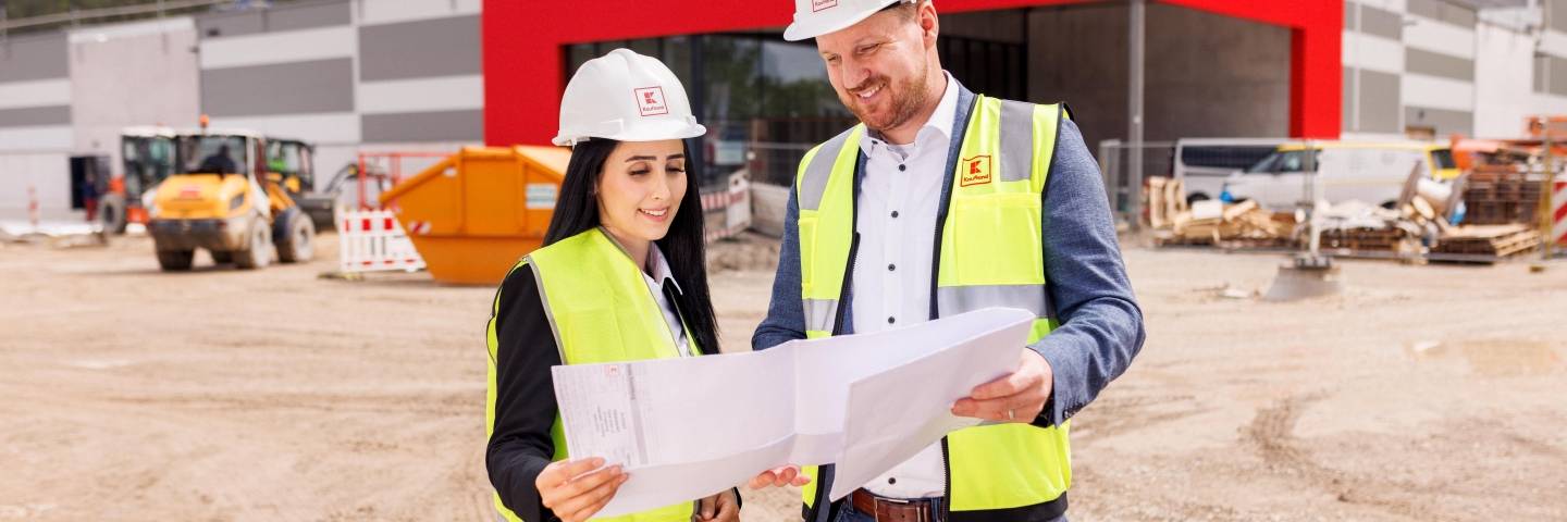 Zwei Mitarbeiter stehen vor dem Bau eines Kaufland-Marktes und sprechen über einen Bauplan, den sie in den Händen halten.