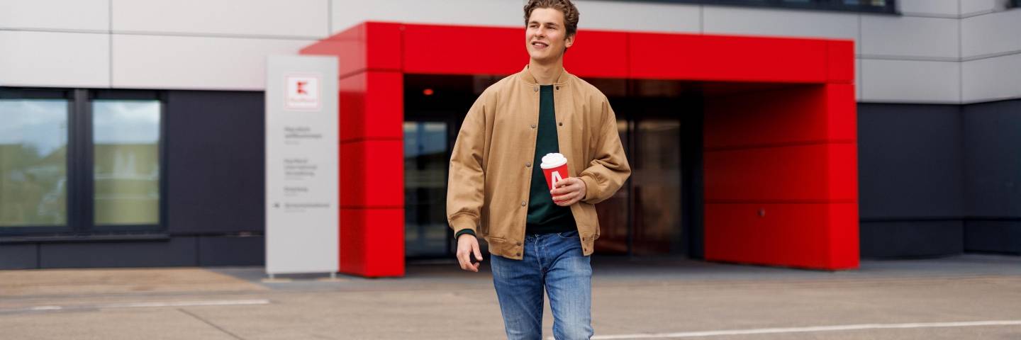 Ein Azubi, ein junger Mann in brauner Jacke, geht über den Platz vor dem Haupteingang der Kaufland-Zentrale.