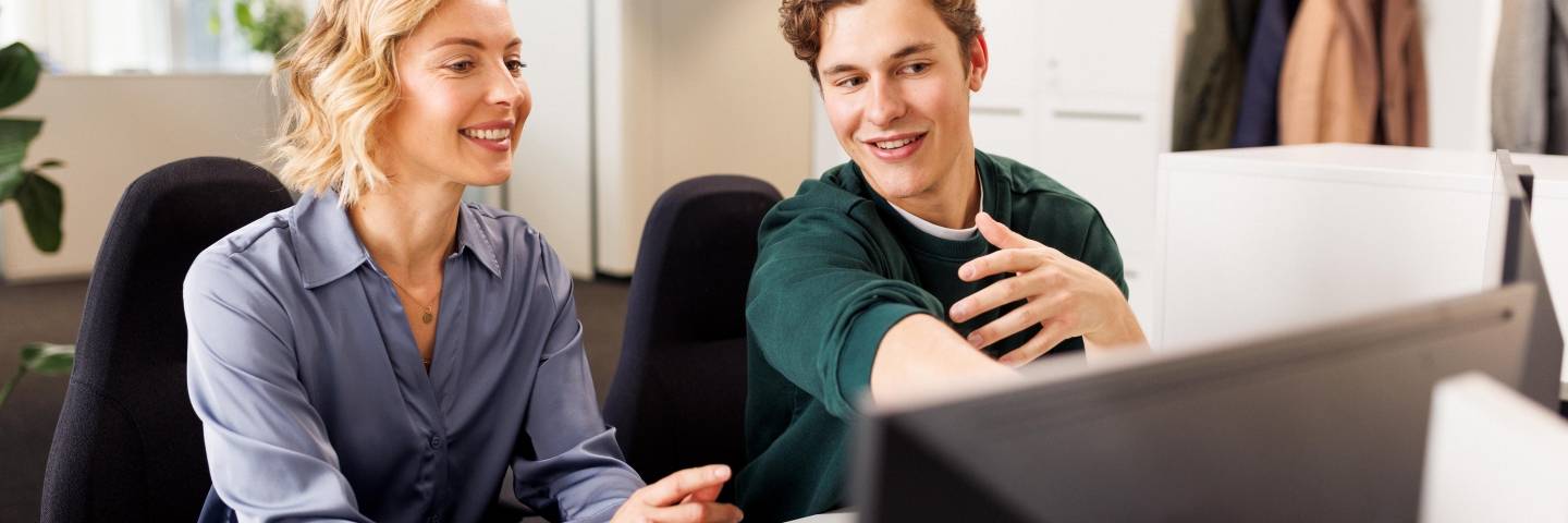 Eine Ausbilderin und ein Student sitzen am Arbeitsplatz vor einem Computer. Die beiden unterhalten sich.
