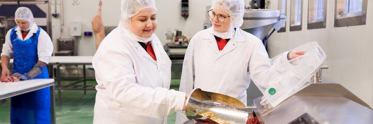 In der Fleischzubereitung stehen ein Azubi und ein Ausbilder in weißer Schutzkleidung vor einem Fleischzerkleinerer. Der Azubi gibt mit einer Kanne Gewürze zum Fleisch.