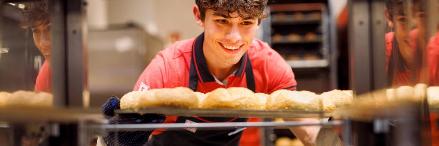 Junger Verkaufsmitarbeiter von Kaufland zieht ein Blech frischgebackener Brötchen aus dem Backregal.