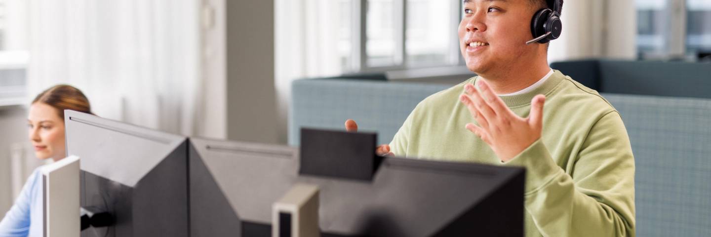 Ein Azubi sitzt mit Headset und im hellgrünen Pullover vor einem Computer. Er führt ein Gespräch.