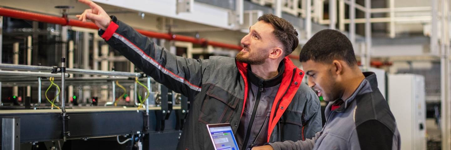 Ein Elektroniker von Kaufland inspiziert mit seinem Kollegen, der einen aufgeklappten Laptop in der Hand hält, eine Logistik-Anlage.