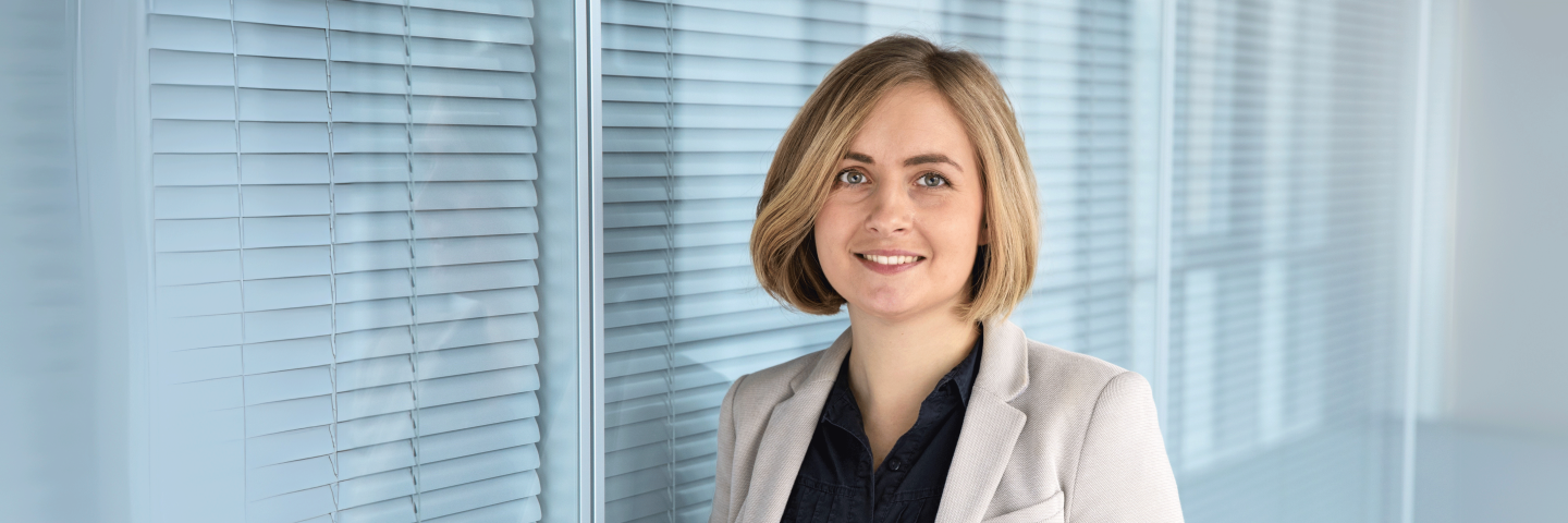 Frau mit blonden Haaren in schwwarzer Bluse und beigem Blazer steht vor einer Glasfront und lächelt freundlich in die Kamera