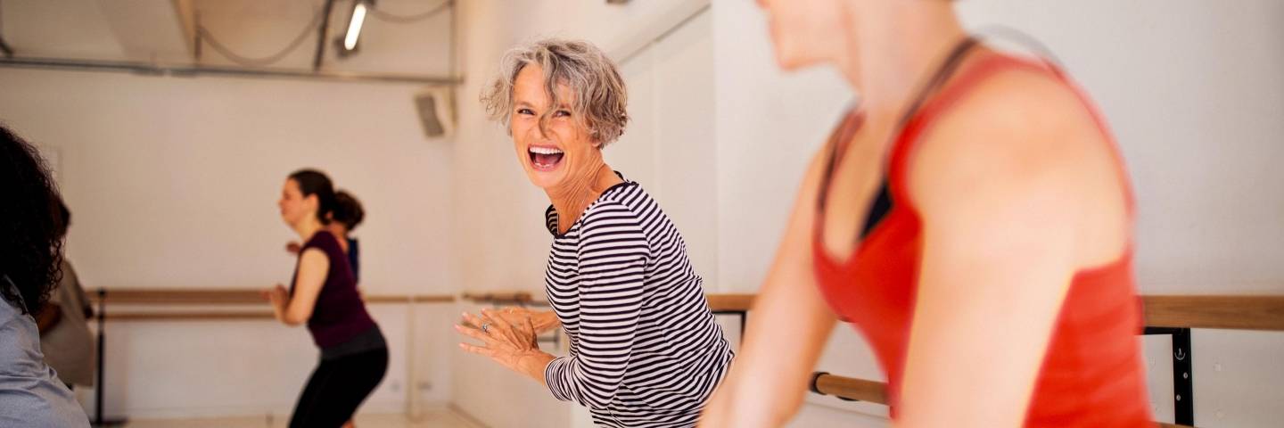 Zwei Frauen nehmen an einem Fitnesskurs teil und lachen dabei. Im Hintergrund sieht man weitere Teilnehmer.