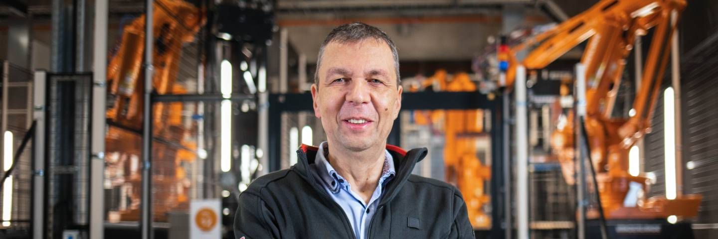 Teamleiter für Automatisierung am Logistikzentrum Geisenfeld steht mit verschränkten Armen vor einer Roboteranlage und lächelt freundlich in die Kamera