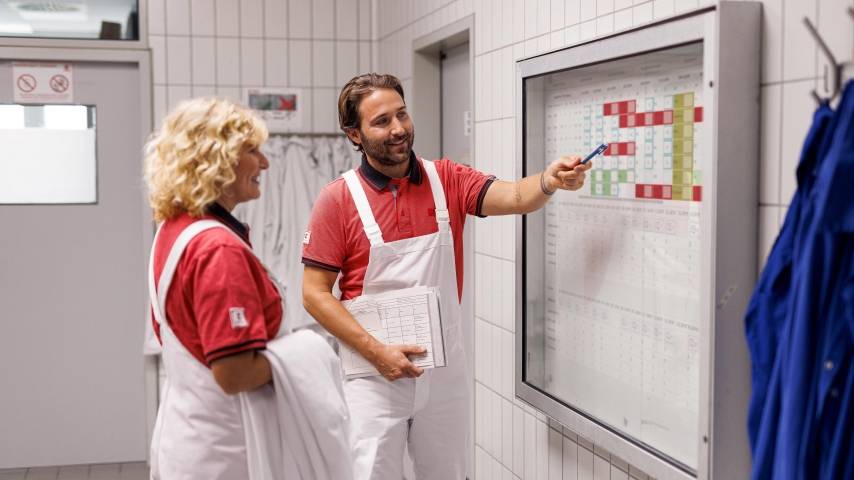 Eine Mitarbeiterin und ein Mitarbeiter in weißer Arbeitskleidung vor einem Schaukasten in einem Kaufland-Fleischwerk.