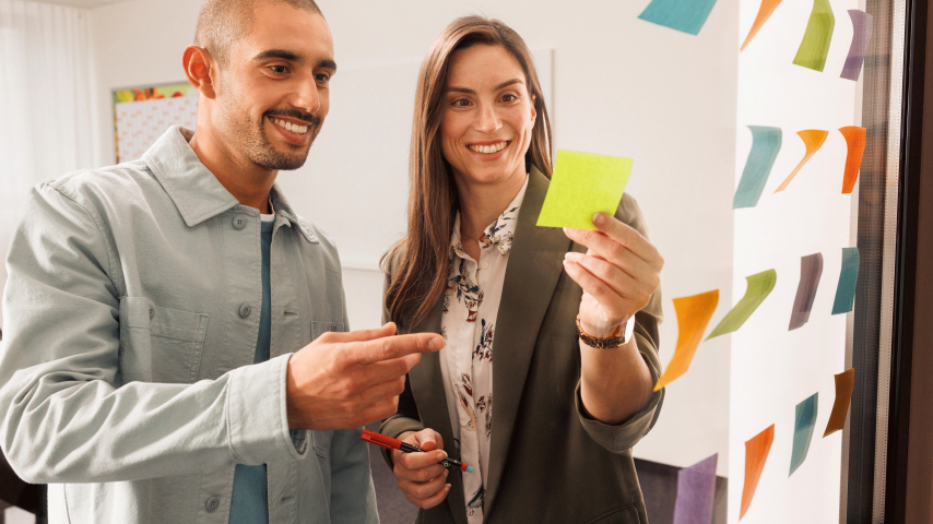 Ein Mitarbeiter und eine Mitarbeiterin befinden sich in einem Meeting. Die beiden stehen vor einer gläsernen Wand und arbeiten mit Post-Its.