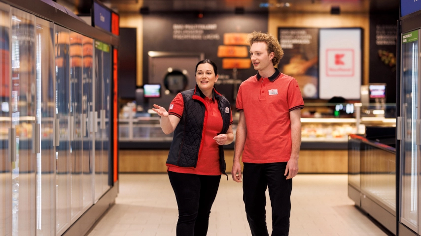 Eine weibliche Führungskraft und ein Mitarbeiter befinden sich auf einem Rundgang durch eine Kaufland-Filiale. Die Führungskraft spricht zum Mitarbeiter.