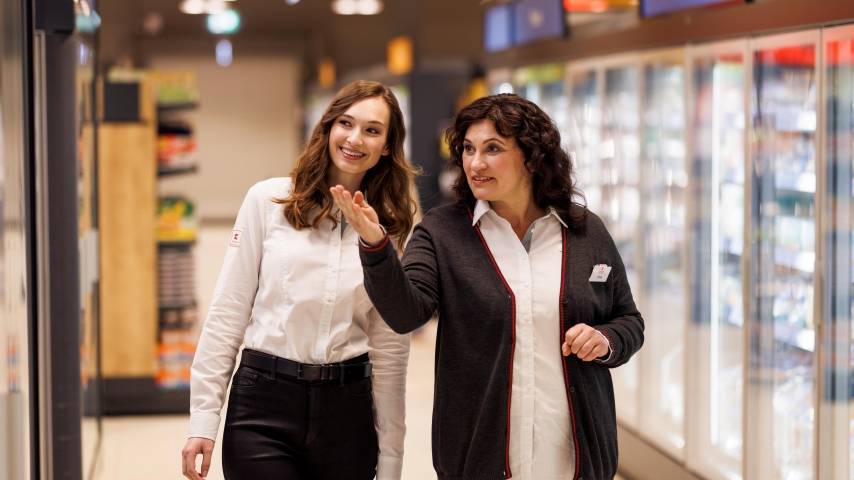 Eine weibliche Führungskraft und eine Mitarbeiterin befinden sich auf einem Rundgang durch eine Kaufland-Filiale. Die beiden sind im Gespräch. Hinter ihnen sind übermannsgroßen Kühlschränken zu sehen.