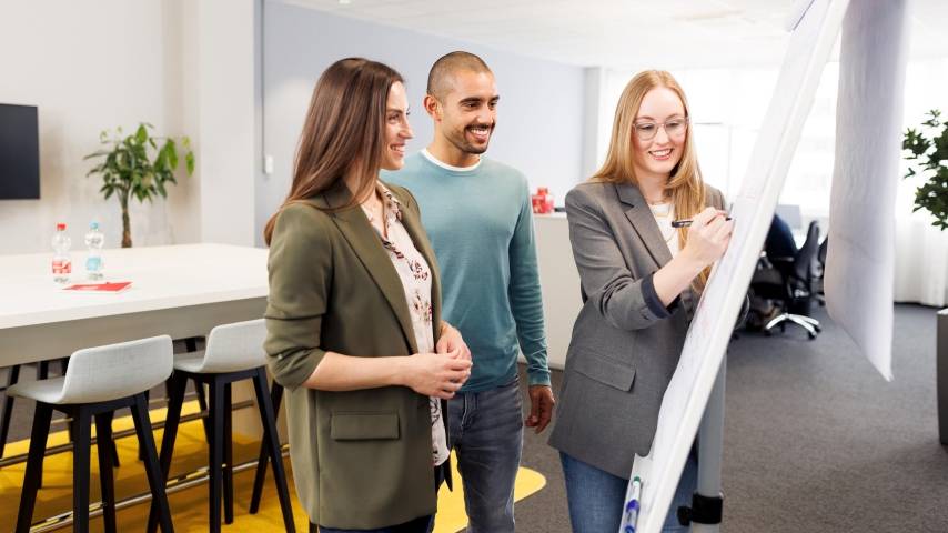 In der Workshopsituation stehen eine junge Frau und ein junger Mann, beide Trainees, mit einer Ausbilderin vor einem Flipchart.