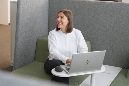 Projektleiterin Patricia aus dem Immobilienbereich sitzt in einer Sitzecke im Büro am Laptop und lacht zur Seite.