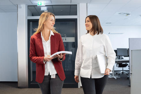 Zwei Frauen laufen einen Gang im Büro entlang und schauen sich nett an.