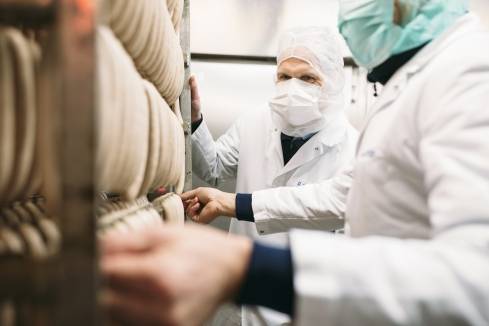 2 Mitarbeiter mit Mundschutz und Schutzkleidung vor einem Regalstand mit aufgehangenen Würsten bei der Qualitätssicherung im Kaufland-Fleischwerk.