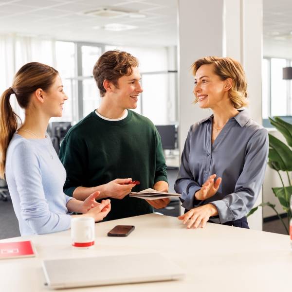 Kollaboratives Arbeiten mit einer weiblichen Führungskraft und zwei Azubis. Die drei stehen in legerer Kleidung am Hochtisch und unterhalten sich.