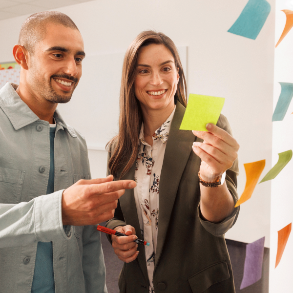 Ein Mitarbeiter und eine Mitarbeiterin befinden sich in einem Meeting. Die beiden stehen vor einer gläsernen Wand und arbeiten mit Post-Its.