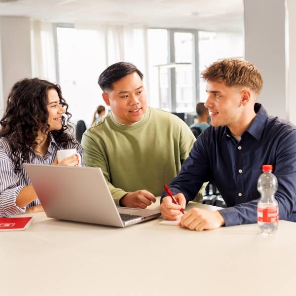 Kollaboratives Arbeiten mit einem Azubi, einem Mitarbeiter und einer Studentin. Die drei jungen Leute stehen in legerer Kleidung am Hochtisch vor einem aufgeklappten Laptop und unterhalten sich.