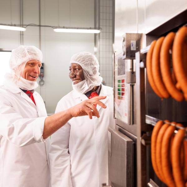 Ein Mitarbeiter und ein Azubi in weißer Schutzkleidung befinden sich in der Rauchkammer und unterhalten sich. Der Mitarbeiter führt eine Qualitätskontrolle durch, während der Azubi eine Maschine bedient. Rechts hängen zwei Reihen von Würsten an Querstangen.