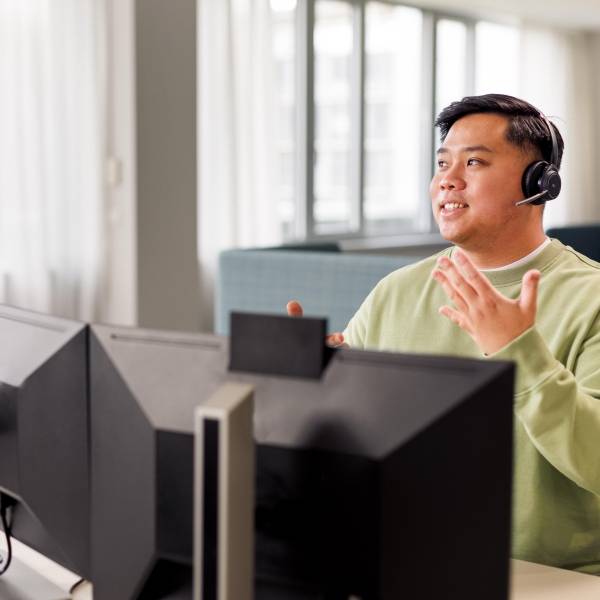 Ein Azubi sitzt mit Headset und im hellgrünen Pullover vor einem Computer. Er führt ein Gespräch.