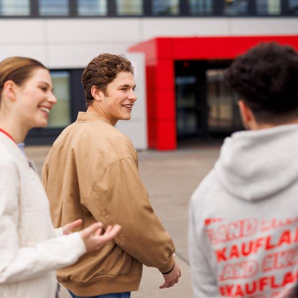 Drei Azubis gehen auf den Haupteingang der Kaufland-Zentrale zu. Links geht eine junge Frau, rechts neben ihr zwei junge Männer. Alle drei sind salopp gekleidet, man sieht sie in seitlicher Ansicht bzw. von hinten.