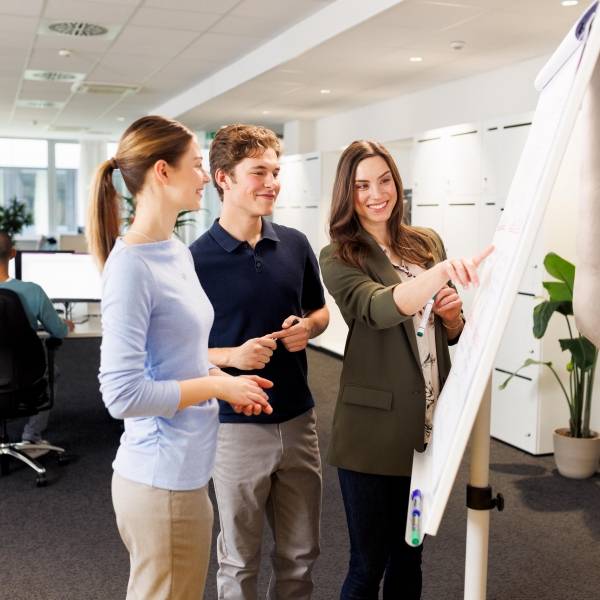 Zwei Azubis, eine junge Frau und ein junger Mann, sowie eine Ausbilderin befinden sich in einer Workshopsituation. Die drei stehen vor einem Flipchart und brainstormen.