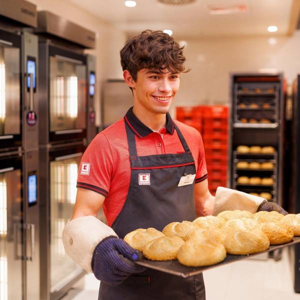 Junger Verkaufsmitarbeiter von Kaufland mit einem Blech frischgebackener Brötchen, die er gleich in die SB-Backwarenregale einsortiert.
