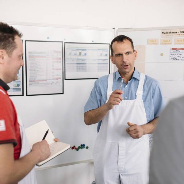 3 Kaufland-Fleischwerk-Mitarbeiter in der Besprechung. Ein Mitarbeiter macht sich Notizen.