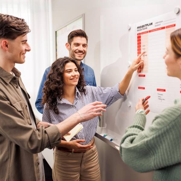 Zwei junge Frauen und zwei junge Männer stehen dicht gruppiert vor einem Projektboard, an dem sie gemeinsam arbeiten.