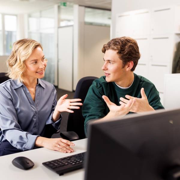 Eine Ausbilderin und ein Student sitzen am Arbeitsplatz vor einem Computer. Die beiden unterhalten sich.