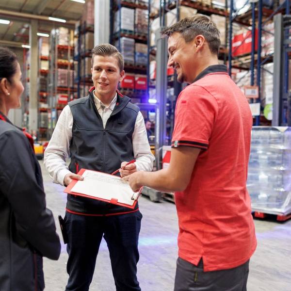 Logistik-Mitarbeiter von Kaufland unterhält sich im Lager mit seiner Kollegin und seinem Kollegen und macht sich Notizen auf seinem Clip-Board.