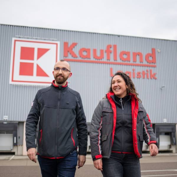 Zwei Logistik-Mitarbeiter bei Kaufland laufen vor einem Logistikzentrum und lachen.