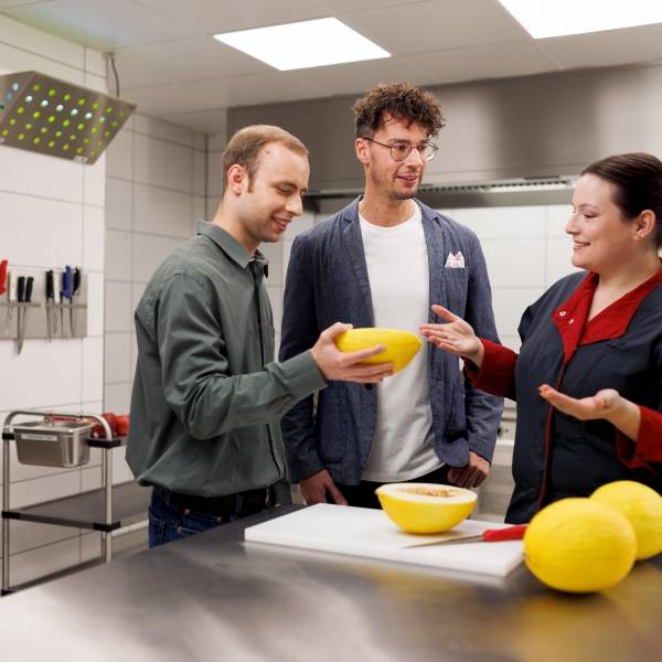 Zwei Mitarbeiter und eine Mitarbeiterin stehen in einer Laborumgebung am Tisch. Sie nehmen eine Verkostung vor und prüfen Ware.