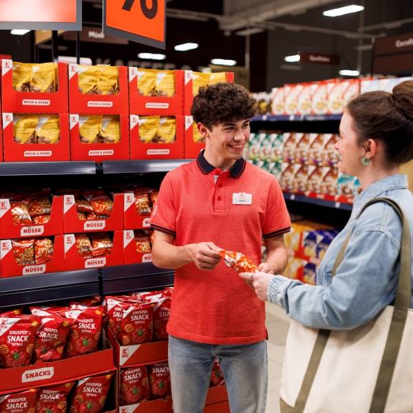 Ein Azubi in rotem Kaufland-T-Shirt und eine Kundin mit umhängender Einkaufstausche stehen vor einem Regal in einer Kaufland-Filiale. Der Azubi erklärt der Kundin, wo sie etwas findet.