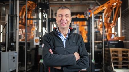 Teamleiter für Automatisierung am Logistikzentrum Geisenfeld steht mit verschränkten Armen vor einer Roboteranlage und lächelt freundlich in die Kamera